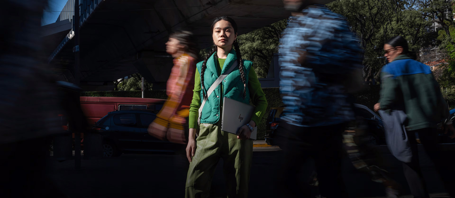 A woman in a green jacket and pants walks on the street, with an HP laptop in her hand.